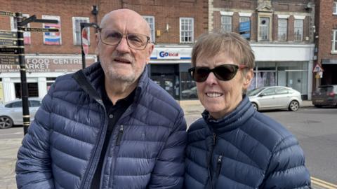 Peter is on the left, he is bald and wearing glasses. he is wearing a navy blue puffer coat. Anne is on his right. she has short brown hair and black sunglasses. She is wearing a navy blue puffer coat.