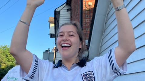 A woman in a white England top cheering, facing the camera,, her arms raised, with a sign reading RIVERSIDE INN hanging outside the upper storey of a brick and white-cladded building. There is a blue sky above.