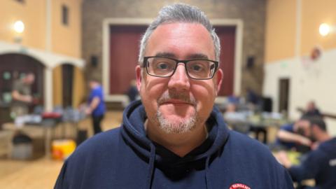 A man wearing black rimmed glasses smirks at the camera - he has grey hair in a quiff and a goatee beard