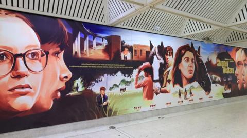 A painted mural featuring the faces of youngsters, in mixture of full face and profile, some next to horses heads. In the background are scenes including a train on a viaduct and a horse under a tree. The artwork's title Stepney Western is written to the far right.