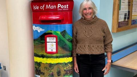 Lesley Sleight smiling at the camera standing next to a large post box decorated in a painted landscape scene. She is wearing a brown jumper and black jeans and it standing in the hospital foyer with a pale blue wall behind her. The design on the red post box features green hills, yellow flowers and a blue sky with one white cloud on the left of the image. The numbers 50 are painted in gold lettering at the bottom of the image.