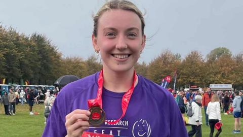 Young white woman standing in the middle of the shot, she is wearing purple r-shirt, and she has a bronze medal, there is a park with people galore behind her.