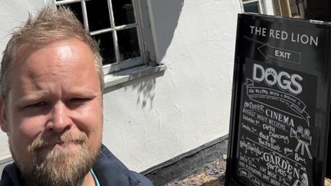 Tom, outside a pub, with a pub sign to the right. He has short fair hair and a beard. There are two windows in the building. 