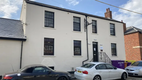 A two-storey whitewash building with large windows with a black door with three cars parked outside it.