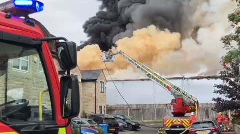 Black, orange and grey smoke rises in a huge plume from a large grey industrial building which sits behind a wooden fence at the end of a residential street. The houses are made of larger beige stone bricks and sit over two storeys. They are all of similar design like so many private housing estates. Lots of cars are parked in front of the houses. The cab of a fire engine is in the foreground. In front of it, a flat bed fire vehicle has a ladder mounted on it. It is extended towards the flames with, presumably, firefighters directing water towards the flames from the cherry picker basket at the end of the ladder. Two firefighters in hi-vis and helmets stand in the distance, backs to shot.