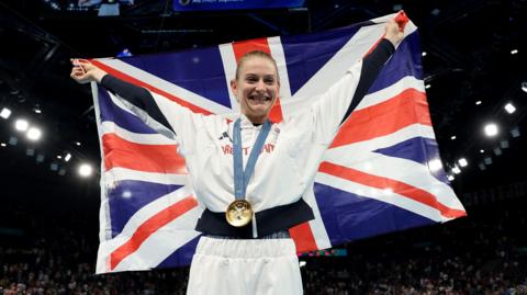 Bryony Page in a Team GB tracksuit looking and smiling at the camera with an Olympic gold medal round her neck. She is holding a Great Britain flag behind her back.