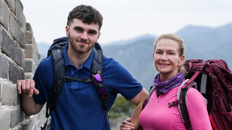 Tom is standing on the right looking at the camera smiling. He has brown hair, and is wearing a blue short sleeved polo top and a black rucksack on his back. Caroline is on the right looking into the camera smiling. She has a pink top on with a purple and white scarf around her neck, and a burgundy rucksack on her back.