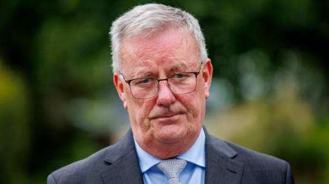 A man is looking into the camera. He has a neutral expression. He's wearing glasses and a grey suit with a blue shirt and a grey tie with a maze design. He's outside. Trees in the background are out of focus. 