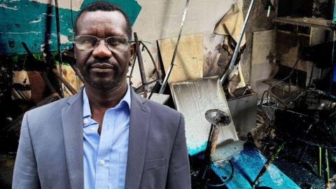 Mohammed Idris has black hair and is wearing glasses, a blue blazer and light blue shirt. In the background is the interior of his cafe after it was burnt. 