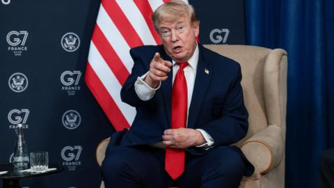 Donald Trump sat in a cream coloured armchair points with his finger. Behind him is a G7 logo and Unites States flag.