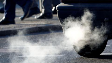 An exhaust pipe of a car is pictured on a street, with white smoke billowing out in a trail. People can be seen walking 