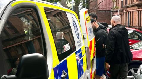 A man is escorted into the back of a blue, white and yellow Police Scotland van by two plain-clothes police officers on a residential street. Cars are parked nearby.