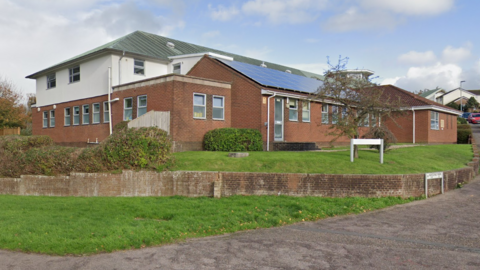 A street-view of Honiton Surgery in Devon. It is a red-brick building with a small wall surrounding it. There is grass and bushes in front of the building and two white street signs on the right. 