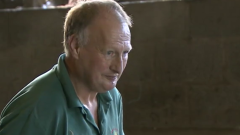 A screenshot from a TV interview with David Knipe. He has short receedeing white hair and blue eyes. He wears a green polo shirt and is inside one of his poultry sheds. He is looking at something out of shot to the side.