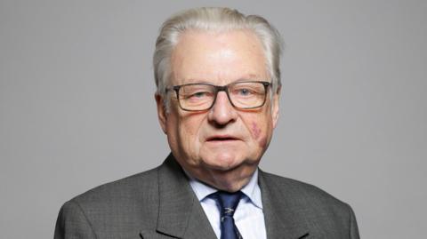 An official House of Lords photograph of Lord Elis-Thomas. He is looking directly at the camera and wearing glasses, a grey suit, light blue shirt and dark blue tie.  