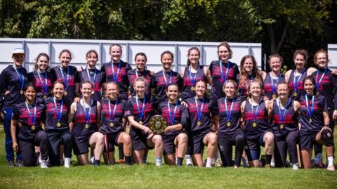 A group shot of the Bristol Women's teammates wearing their black and red uniform. There are two rows of people, with those in the forefront down on one knee. They are all smiling at the camera with their arms around each other, and two women in the front are holding a wooden trophy plaque. 