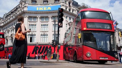A red London bus turns right as a pedestrian passes by on Oxford Street in central London.