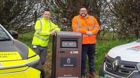 A picture of two people stood either side of the talking bin. It is brown in colour and there is a vehicle either side of the bin.