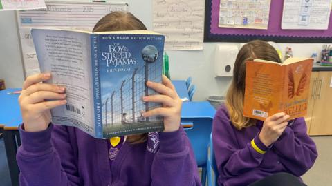 Two schoolgirls hold books up to their faces. You cannot make out what they look like.