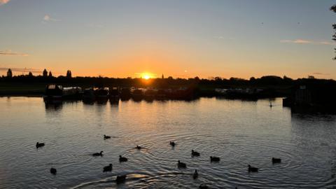 Several ducks sit on a lake in Oxford with a silhouetted horizon in the background under a low orange sun