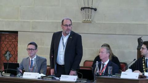 A man wearing a white shirt, black jacket and lanyard is standing up in a council chamber, speaking into a microphone. He is sat next to several other men wearing suits.