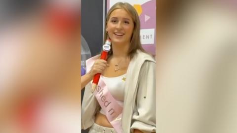 A girl wearing a pink sash and beige jumper smiles at the camera.
