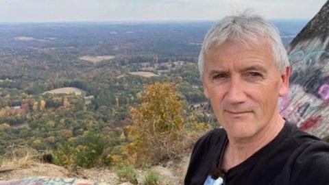 A man with grey hair and black t-shirt takes a selfie on a mountain overlooking lots of trees. 