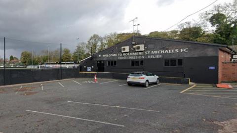 A football clubhouse with WELCOME TO BOLDMERE ST MICHAELS FC written in capital letters across the front. There is a white car parked in front of the clubhouse.