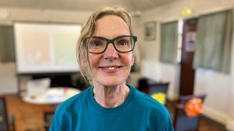 A women with short blonde hair, green rimmed glasses and a blue t-shirt. She is looking just above the camera and smiling. 