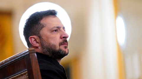 Close up of Volodymyr Zelensky in black blazer looking to the left, a large light illuminating him from behind