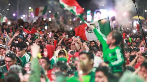 A huge crowd of fans wearing Mexico's green football kit and waving the country's flag celebrate the team's 2-0 World Cup win against Ecuador in Mexico City at night.
