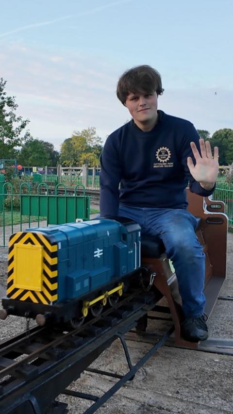 a man sits on a small train and waves at the camera.