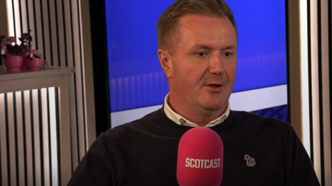 Andrew Murray is sitting in front of a red mic that says Scotcast in white. He is looking off camera and is in mid sentence. He is wearing a dark jumper with a light-coloured shirt underneath it. There are plants behind him.