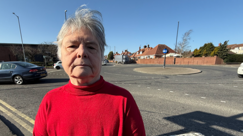 A woman with grey hair and wearing a red-coloured turtle neck jumper stands in front of a roundabout, which has a bricked centre and a black lamppost standing in the middle of it.