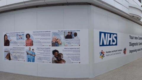 A photo of the exterior of a community diagnostic centre. It shows white walls and banners with photos of people, writing and a large NHS logo. 