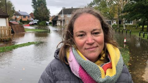 Vhari Russell has shoulder length brown hair, a grey coat with colourful woollen scarf and is standing in front of a flooded housing estate.