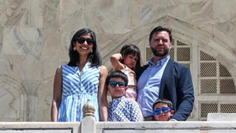 Usha Vance and JD Vance stand with their three kids at the Taj Mahal. JD is holding their daughter. Usha and their two sons wear sunglasses.