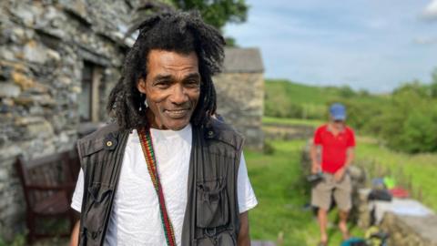 Easton Christian, a man with dreadlocks stands smiling outside a stone cottage as another man stands in the distance by a low stone wall