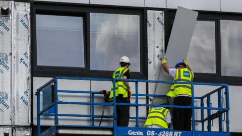 Three workers in hard hats and hi-vis jackets remove a panel of cladding from a building