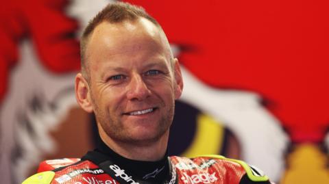 Shane Byrne in a red motorcycle outfit. He is looking at the camera and is smiling. There is red motorsport branding on a wall behind him.