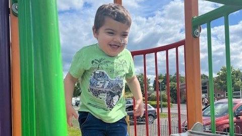 Gunner walking on climbing equipment in the park. He wears a green t-shirt and blue trousers and is smiling. There are parked cars behind him. 