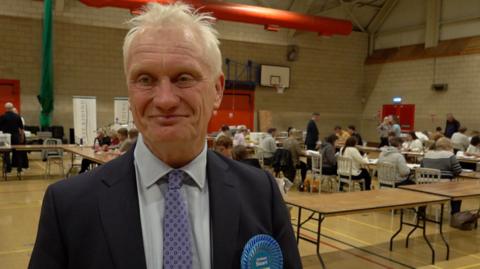 MP Graham Stuart smiles at the election count. He is wearing a Conservative Party rosette. 