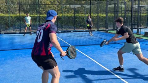There are four men playing padel. On the one side, one of the men has a lime green t-shirt and black shorts with a yellow racquet. The other man is wearing a black t-shirt and black shorts. In the front, there is a man with his back to camera wearing a blue backwards cap, a purple, pink and navy shirt with 'Tulloch 70' on the back and black shorts. He is holding a black raquet. On the right, at the front, there is a man with a black t-shirt, cyan shorts and black shoes. He is hitting the ball.