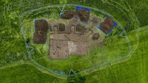 An aerial shot of the dig site. One main area is excavated in a rough square shape, with a series of smaller, rounder dig sites around it. 