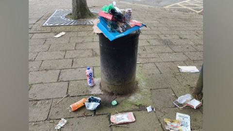 There is a tall black bin which is overflowing with litter. On the floor is rubbish including a Yazoo bottle, an orange boost bottle, a gregs wrapper and a haribo goldbears wrapper. On top of the bin, there is rubbish piled on top of a big Dominos box.