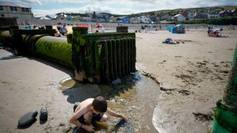 A boy plays in a stream in front of a discharge pipe on a sunny beach in Wales, with sand and sunbathers in the background. 