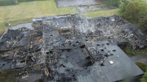 An aerial view of a building that has been destroyed in a fire. The building is covered in ash and soot and some smoke is rising from the site.