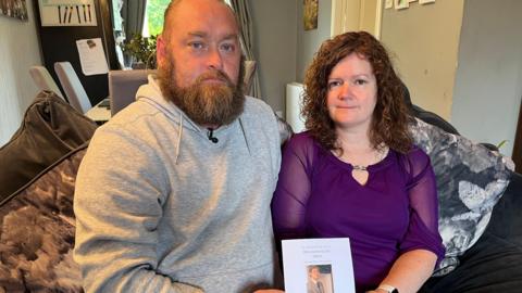 A man and woman sitting next to each other on a sofa in a living room. They are holding an order of service from a funeral