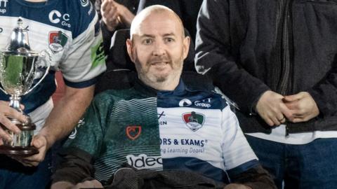 Matt Gallagher sat in an electric wheelchair after a rugby game, posing with the team and his family