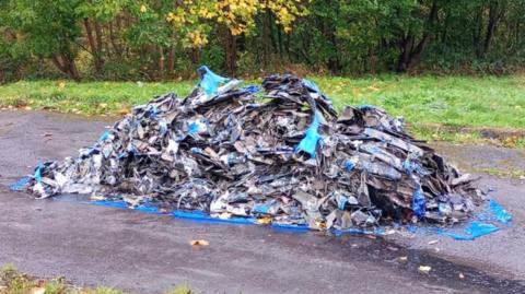 A large pile of rubbish placed on a blue tarp that lies on an asphalt surface. The debris includes a mix of materials such as metal fragments, plastic items, and other refuse. This scene is set outdoors in a park-like area, with green grass and trees visible in the background. Some of the trees have yellowing leaves. The ground surrounding the pile appears wet.